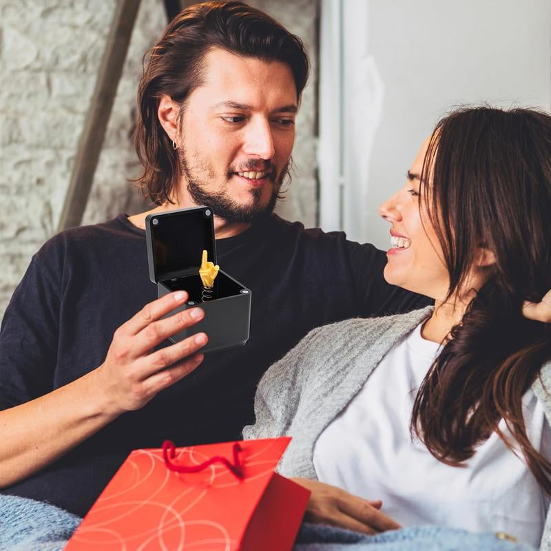 Man gifting woman a black box with a golden middle finger ring, both smiling indoors